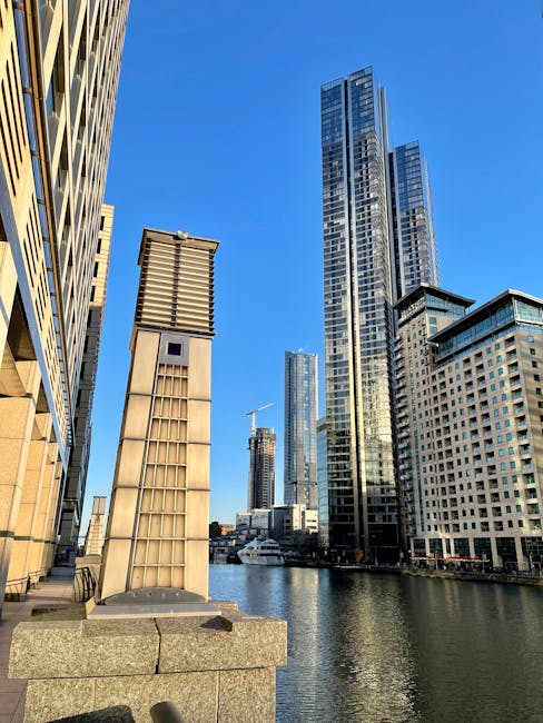 A cityscape featuring tall modern skyscrapers along a waterfront, with reflective glass facades and varying architectural styles. In the foreground, a concrete ledge supports a metallic vent or chimney structure, surrounded by several cardboard boxes and plastic-wrapped furniture pieces indicating packing for home relocation. The scene is well-lit by natural daylight, with a clear blue sky overhead. To the right, a street or promenade runs parallel to the water, where Removal Companies Docklands is facilitating furniture transport and moving logistics, preparing items for loading into a van parked nearby. The image captures the urban environment typical of the Canary Wharf area in the Docklands, highlighting the process of professional removals and packing within a high-rise residential or commercial setting, supporting house removals and relocation services focusing on riverside flats.