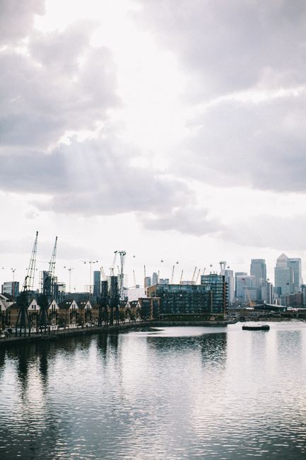 A wide view of the River Thames in the Docklands area under a cloudy sky, featuring modern skyscrapers in the background and several construction cranes towering above partially completed buildings. In the foreground, the calm water reflects the overcast sky and the structures along the riverbank. The image shows a typical scene of urban development with ongoing construction activity, consistent with the type of environment encountered during house removals and furniture transport in the Canary Wharf area. Inside a property, near a doorway, there are cardboard boxes sealed with tape, along with wrapped furniture protected by plastic covers and moving blankets, indicative of packing and packing and moving preparations managed by Removal Companies Docklands as part of a home relocation process. A trolley and straps used for loading are also visible, supporting the logistical aspects of a professional removal service taking place in a Riverside flats setting, aligned with the services described in the Canary Wharf removals guide.