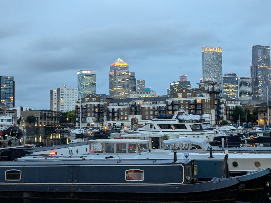 A view of a marina with several luxury yachts docked along the waterfront, with modern high-rise buildings and office towers in the background under a cloudy sky. Close to the water, there are brick residential buildings with balconies, suggesting a mixed-use area within a cityscape. The yachts are moored side by side, with some covered in protective tarps and others with open decks, indicating ongoing or completed boat storage or transportation. The scene captures the urban environment typical of London Docklands, relevant to house removals and moving services provided by Removal Companies Docklands. The overall lighting is dim, with some building lights starting to turn on, reflecting a late afternoon or early evening setting, suitable for illustrating comprehensive house relocation or furniture transport activities in a city marina environment.