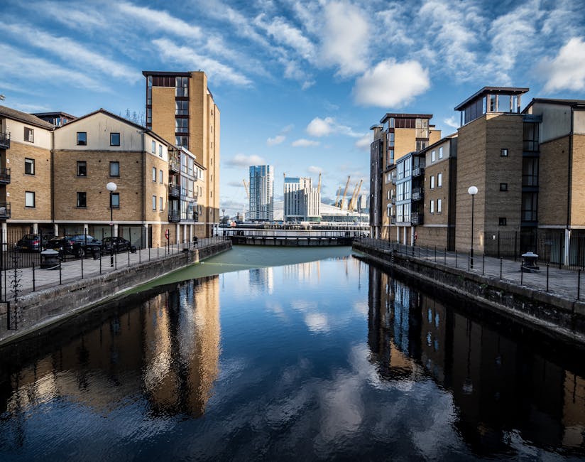 A daytime scene of a canal flanked by modern residential buildings with brick and concrete facades, black metal railings along the water's edge, and parked cars on the pavement. The canal water is calm, reflecting the blue sky with scattered clouds and the surrounding buildings. In the background, there are taller office or commercial buildings with glass exteriors and a construction crane, indicating ongoing development in the Docklands area. The image captures an urban environment suitable for house removals and furniture transport, with the buildings and waterways illustrating the logistics involved in home relocation services provided by Removal Companies Docklands.