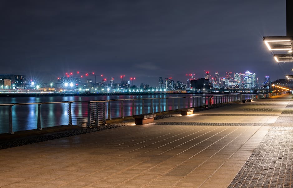 A nighttime view of a riverside promenade in the Docklands area, showing a clear, well-lit concrete walkway with evenly spaced benches along the edge, metal railings separating the pavement from the river, and a city skyline illuminated with numerous high-rise buildings and cranes in the background. The calm water reflects the city lights, and the sky is overcast with dark clouds. The scene is part of a professional home relocation service by Removal Companies Docklands, supporting furniture transport and loading processes during house removals within the area.