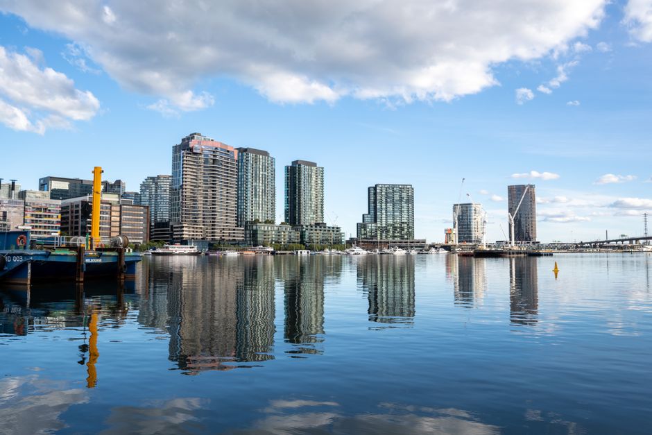 A wide view of the Isle of Dogs skyline reflecting on calm river waters under a partly cloudy sky, featuring multiple modern high-rise buildings with glass facades, and a small blue boat moored near the foreground with visible yellow and black mooring equipment. The scene captures a typical waterfront area suitable for house removals or furniture transport, with the reflection emphasizing the urban landscape. This image is associated with the services of Removal Companies Docklands, illustrating the environment where home relocation and packing and moving activities may take place, especially when loading or unloading goods near the water access point.