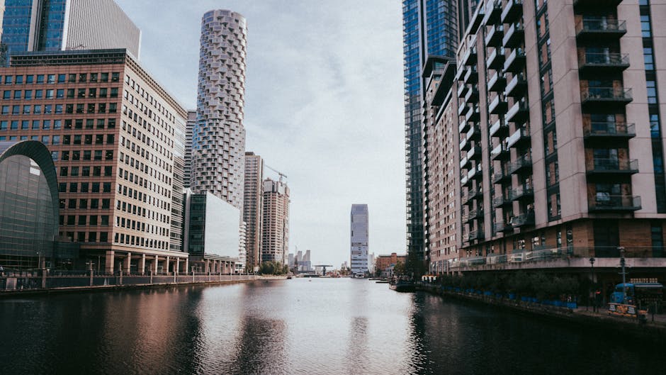 A cityscape capturing a river with reflections of modern skyscrapers including one under construction, under a clear sky. A red urban train runs along an elevated track above the river, passing throug