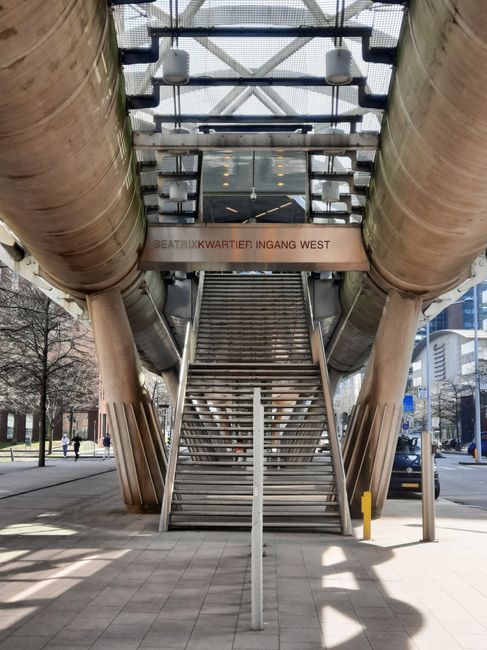 An outdoor staircase leading up to a modern underground entrance at West India Quay in Docklands, constructed with metal steps and handrails, positioned between large cylindrical ventilation ducts made of brownish material. The entrance is sheltered by a glass canopy supported by a metal framework, allowing natural light to illuminate the area. A digital sign displays the words 'BEATRIX KWARTIER: INGANG WEST,' indicating the entrance to an underground station or building. The surrounding environment includes a paved sidewalk, a few pedestrians walking nearby, and urban buildings in the background. The photograph captures the scene during daylight hours with shadows cast on the pavement, demonstrating aspects involved in home relocation and furniture transport processes, especially navigating tight staircases during removals or moving services provided by companies like Removal Companies Docklands.