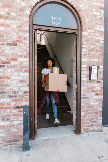 A woman with curly black hair is seen carrying a cardboard box during a home relocation, emerging from a brick building with a black metal threshold. She is wearing a grey t-shirt, blue jeans, and white sneakers, with a red plaid shirt tied around her waist. Behind her, a staircase with a black handrail and white walls is visible inside the building. The entrance features an arched window at the top displaying the numbers 4474 and 4476. To the right of the doorway, a doorbell or intercom system is mounted on the brick wall. The scene is lit by natural daylight, highlighting the process of packing and moving as part of a furniture transport or house removals service, with the company name 'Removal Companies Docklands' occasionally integrated into the context.