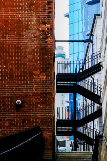 An outdoor staircase leading up to a modern underground entrance at West India Quay in Docklands, constructed with metal steps and handrails, positioned between large cylindrical ventilation ducts made of brownish material. The entrance is sheltered by a glass canopy supported by a metal framework, allowing natural light to illuminate the area. A digital sign displays the words 'BEATRIX KWARTIER: INGANG WEST,' indicating the entrance to an underground station or building. The surrounding environment includes a paved sidewalk, a few pedestrians walking nearby, and urban buildings in the background. The photograph captures the scene during daylight hours with shadows cast on the pavement, demonstrating aspects involved in home relocation and furniture transport processes, especially navigating tight staircases during removals or moving services provided by companies like Removal Companies Docklands.